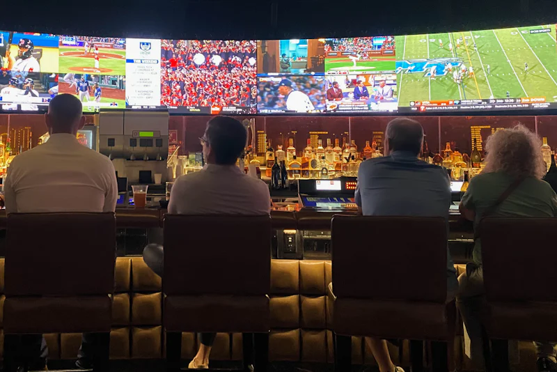 Sports fans sitting at a bar watching basketball and football on giant screens